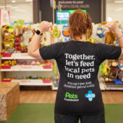 A Pets at Home colleague facing away from the camera in store as she's pointing to the back of her shirt that says: Together, let's feed local pets in need. Pick up a pet food donation parcel today. Pets Foundation and Blue Cross logos below the text.