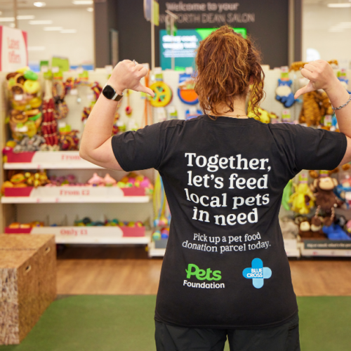 A Pets at Home colleague facing away from the camera in store as she's pointing to the back of her shirt that says: Together, let's feed local pets in need. Pick up a pet food donation parcel today. Pets Foundation and Blue Cross logos below the text.