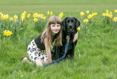 A girl and a black assistance dog in a green field with yellow flowers. The girl has her arm around the dog.