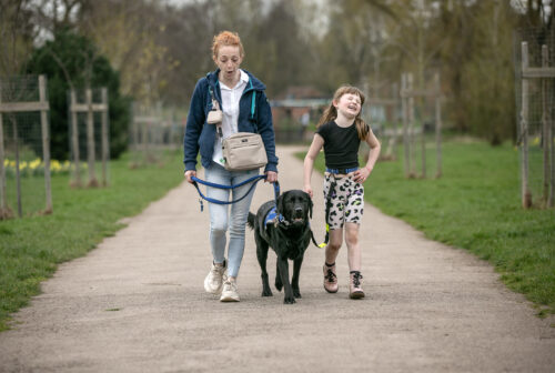 A girl and a woman walking with a black assistance dog. 