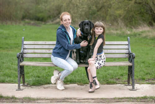 A woman and a young girl hugging a black assistance dog, all sitting on a bench.